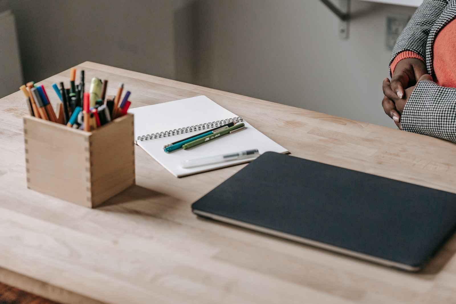 A wooden box of pens and pencils, a spiral notebook and a closed laptop sit on a pale wood desk.