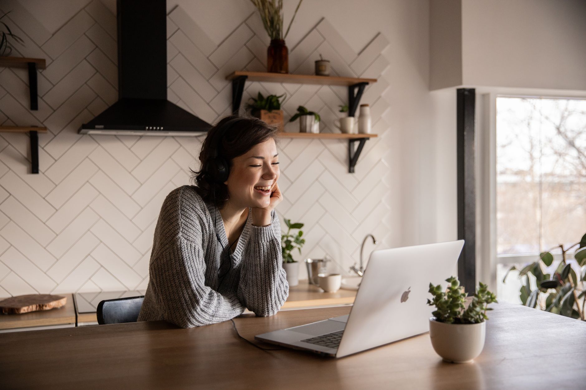 A woman in headphones laughing at a laptop at a kitchen counter
