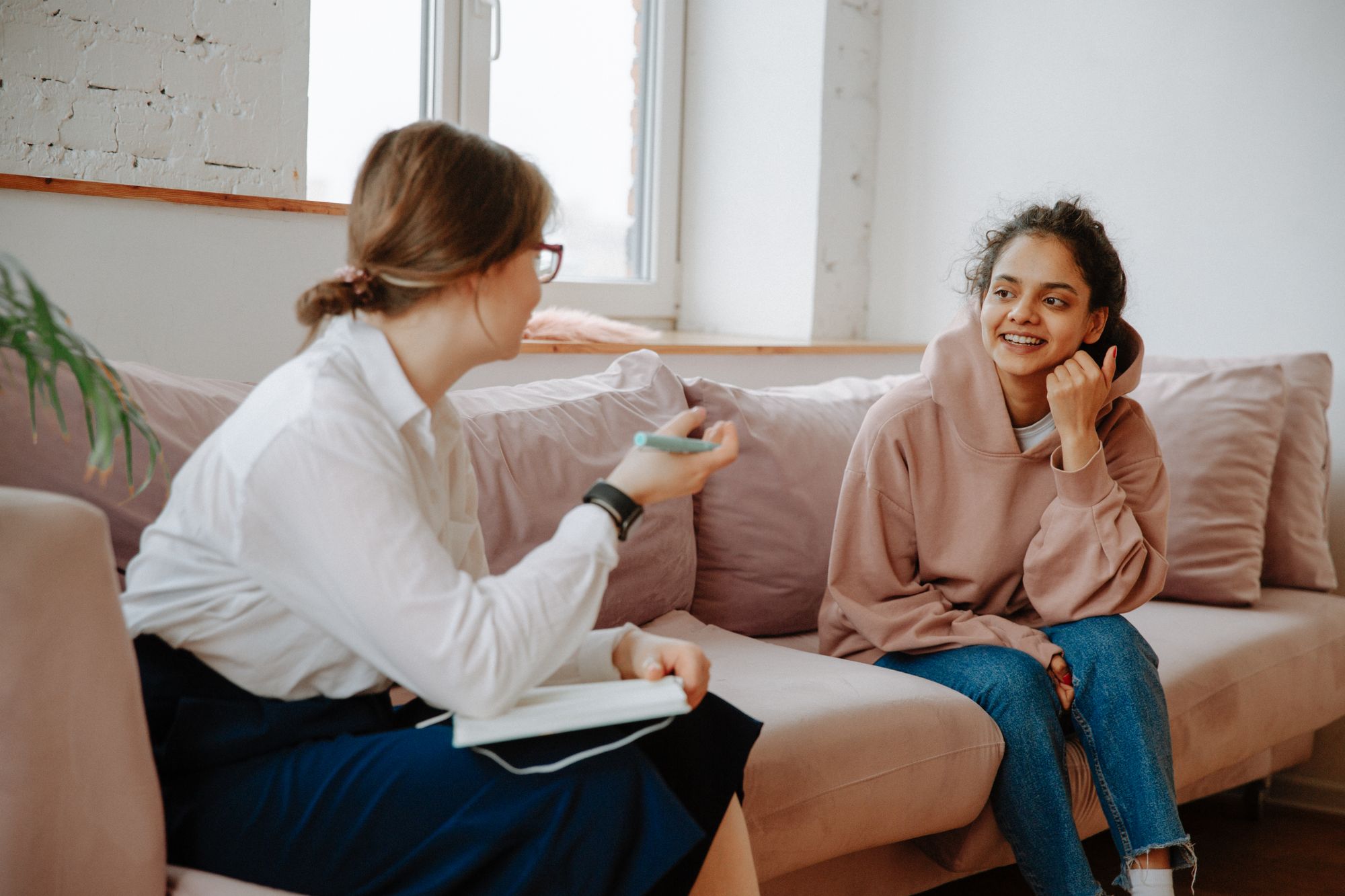 Two women talking on a pink sofa, one holding a notebook