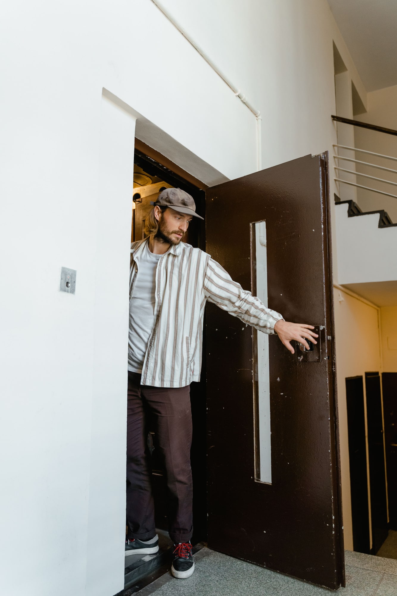 A man in a cap and a striped shirt steps through a doorway, holding a dark metal door open
