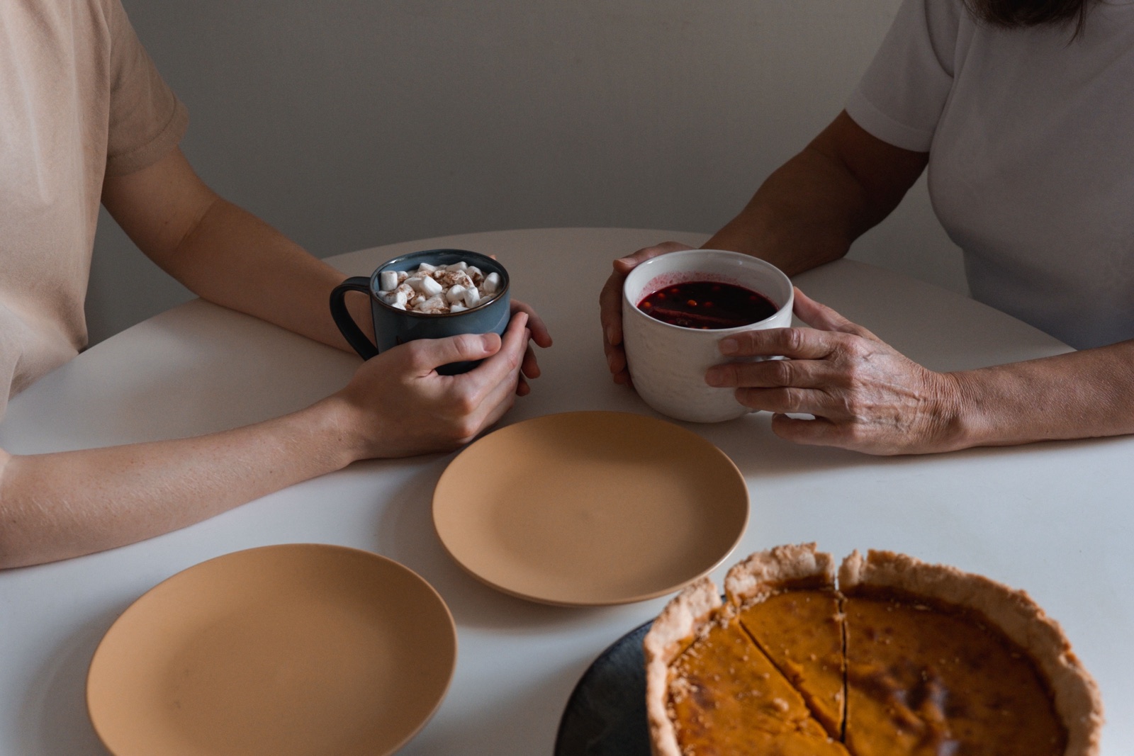 Two people sit across a white table, each holding a mug, with two empty plates and a pumpkin pie in front of them.