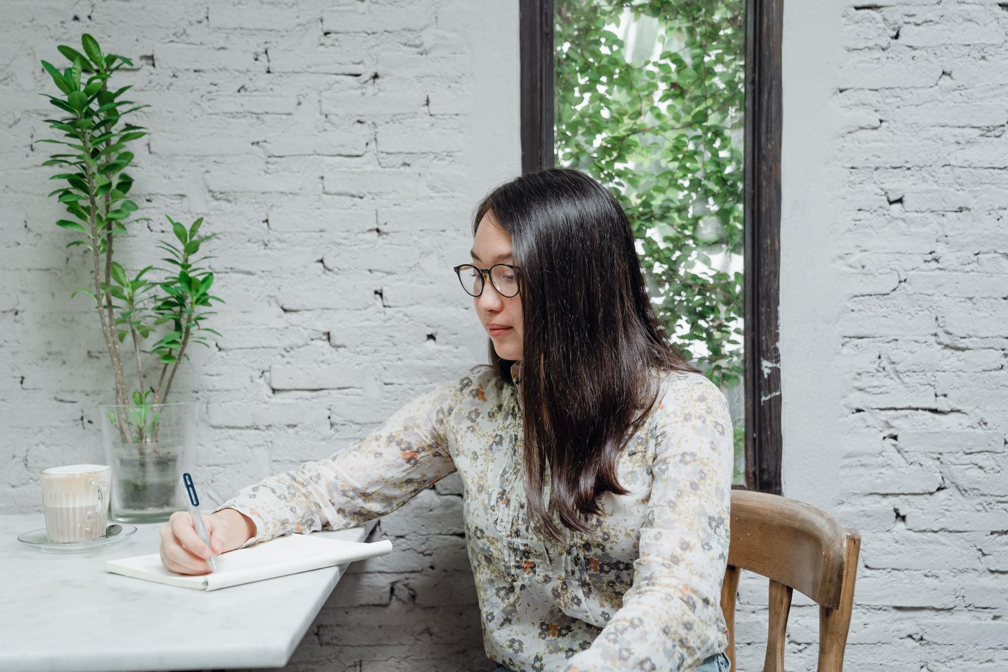 A woman writing in a notebook at a table by a window