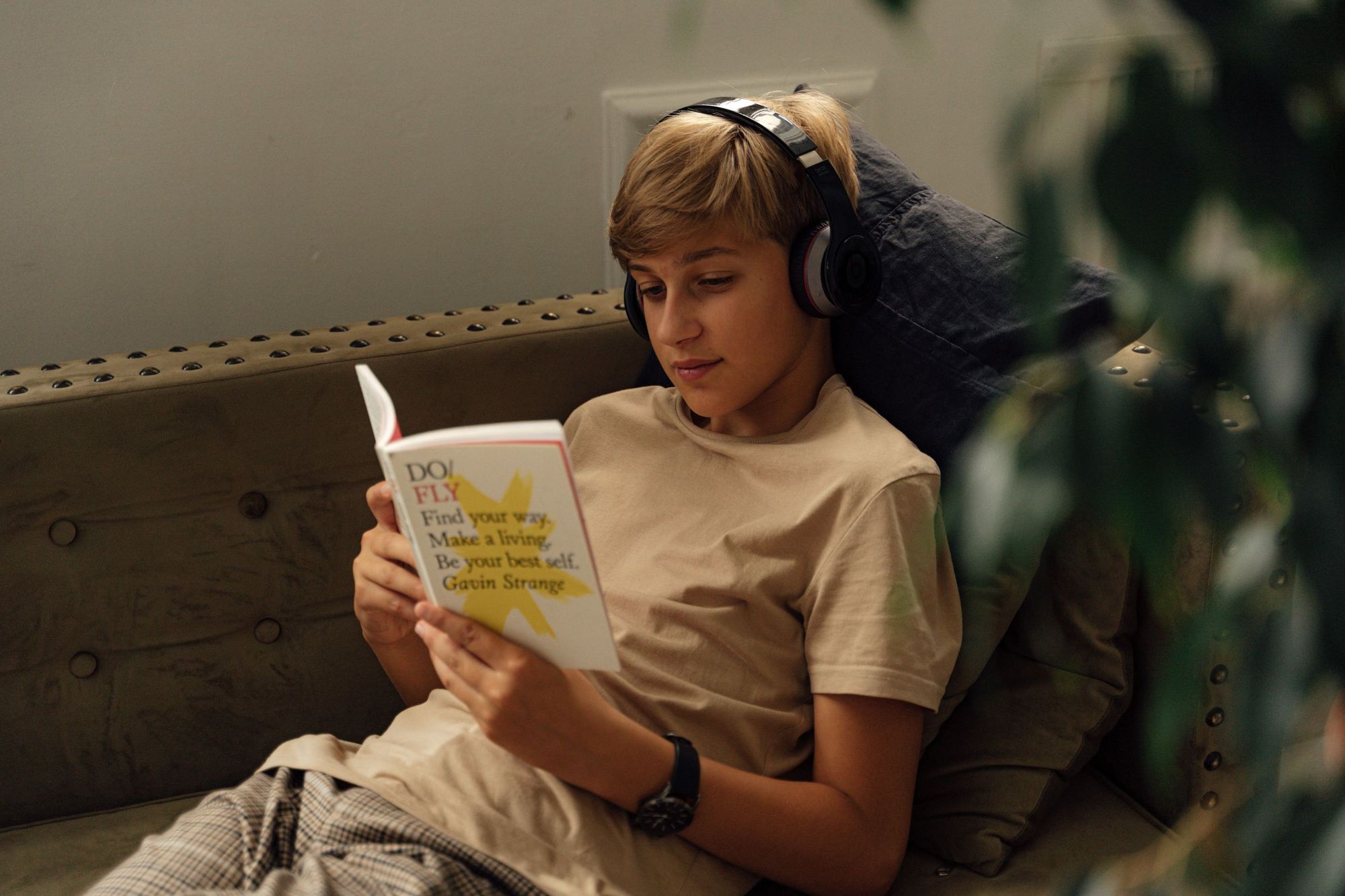 A boy wearing headphones reading a book on the couch