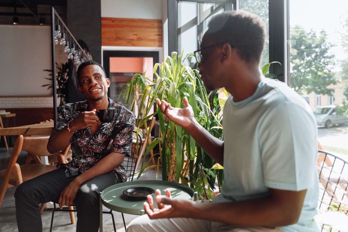 Two men talk at a small café table beside a large window, one smiling and holding a cup.