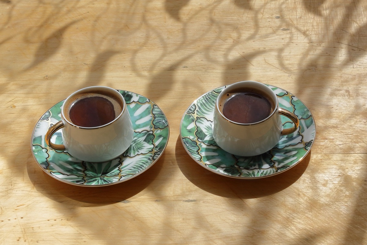 Two cups of coffee on patterned saucers sit side by side on a sunlit wooden table.