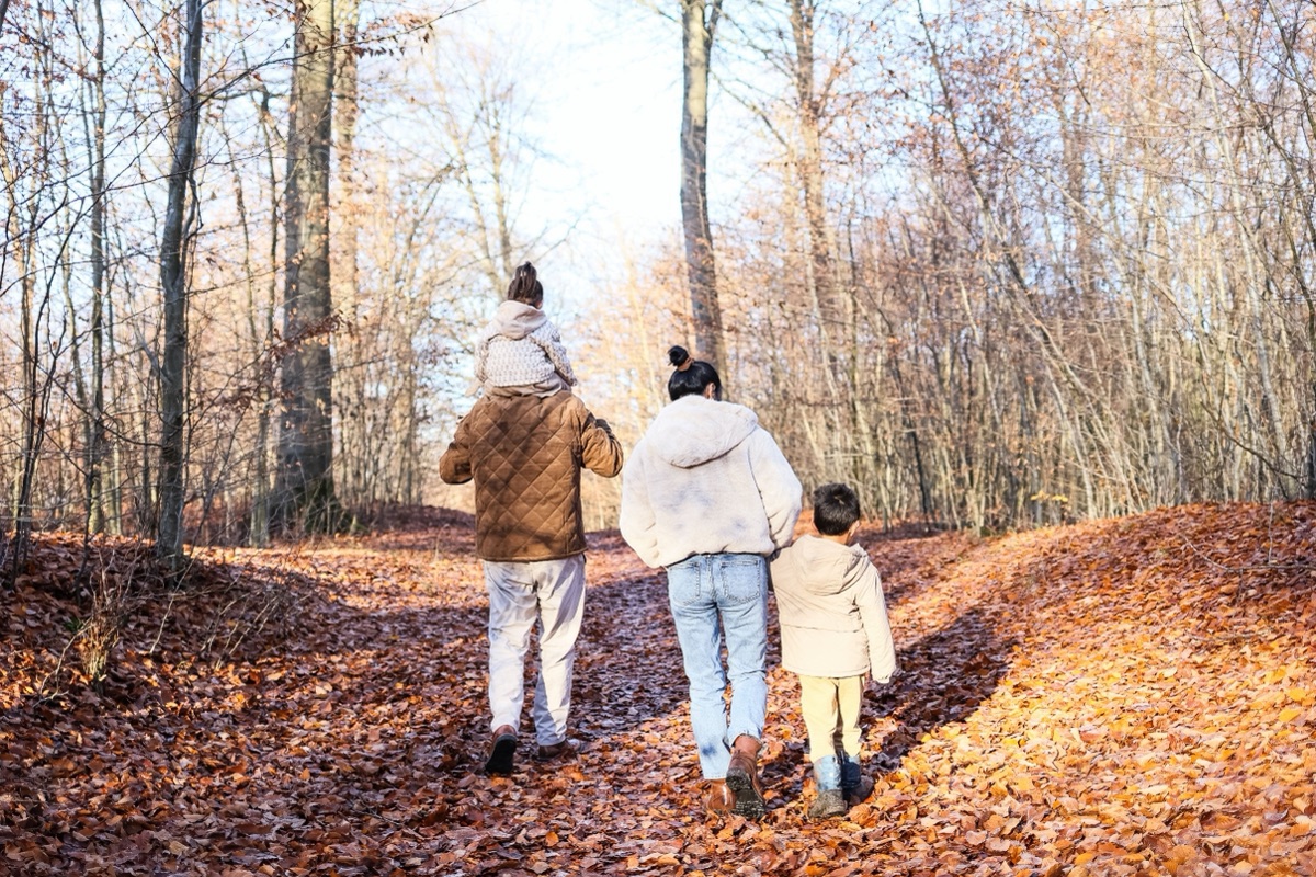 A family seen from behind walks through a leafy autumn forest, one child riding on shoulders.