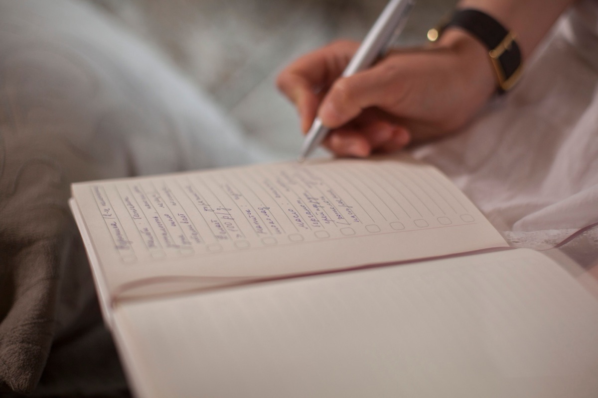 A hand with a wristwatch writes a list in an open lined notebook.
