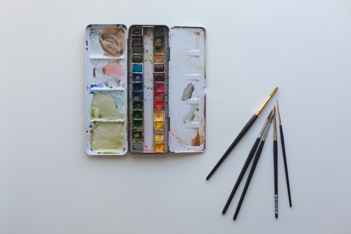 A well-used tin of watercolour paints lies open beside five fine brushes on a white table.