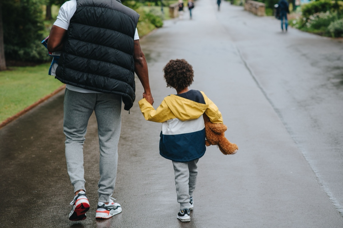 An adult and a small child carrying a teddy bear walk hand in hand along a wet path, seen from behind.