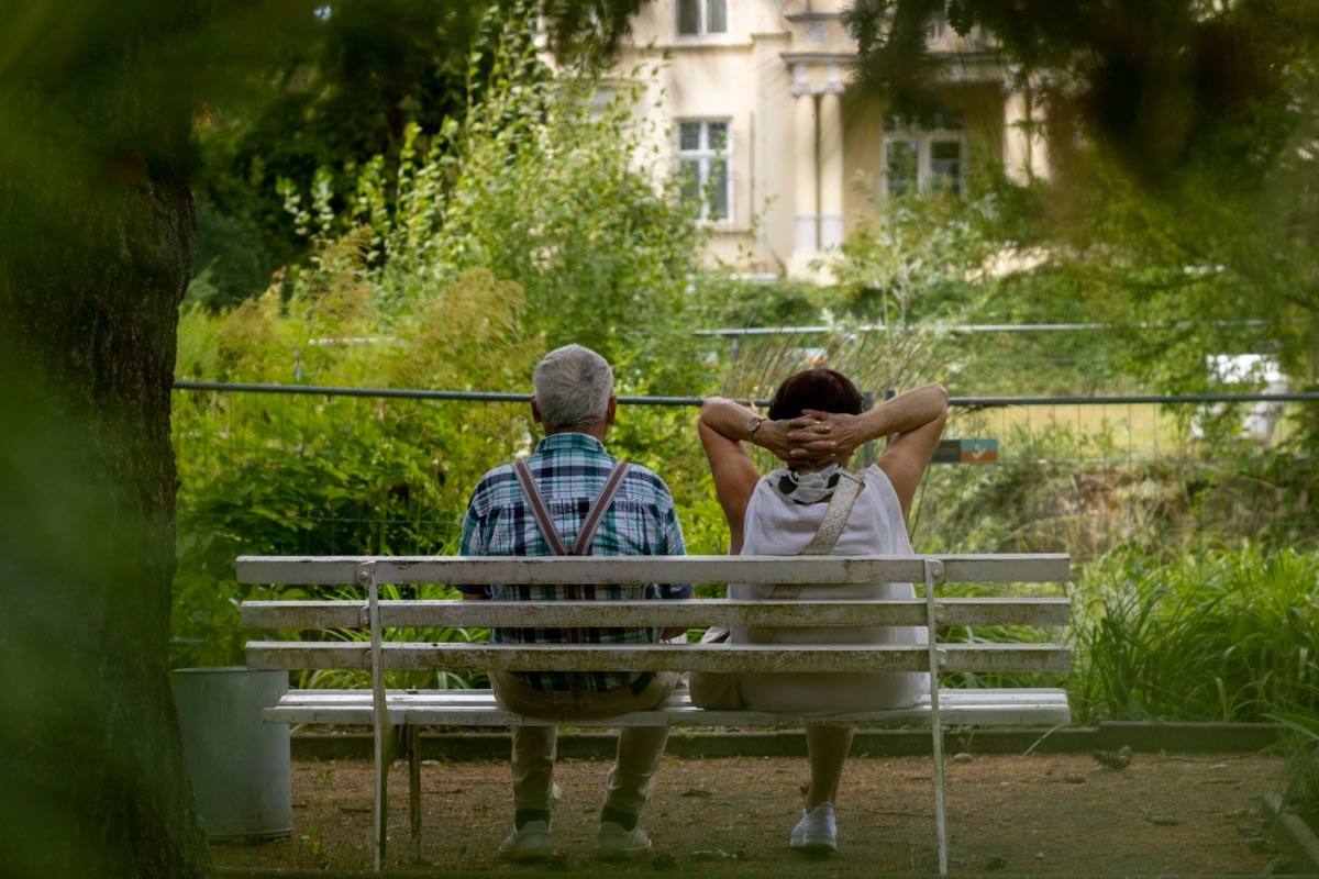 Two older adults sit side by side on a white park bench, seen from behind, facing a garden.