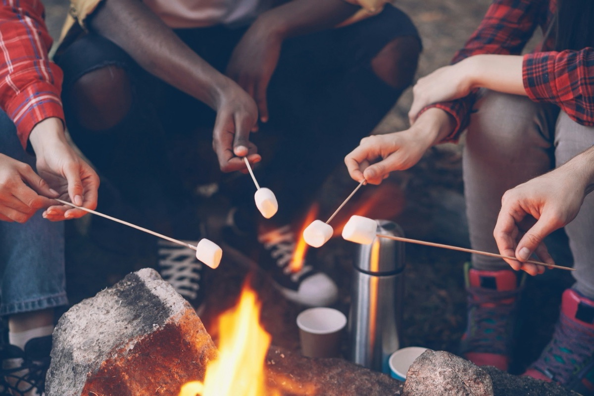Several hands hold sticks with marshmallows over a small campfire.