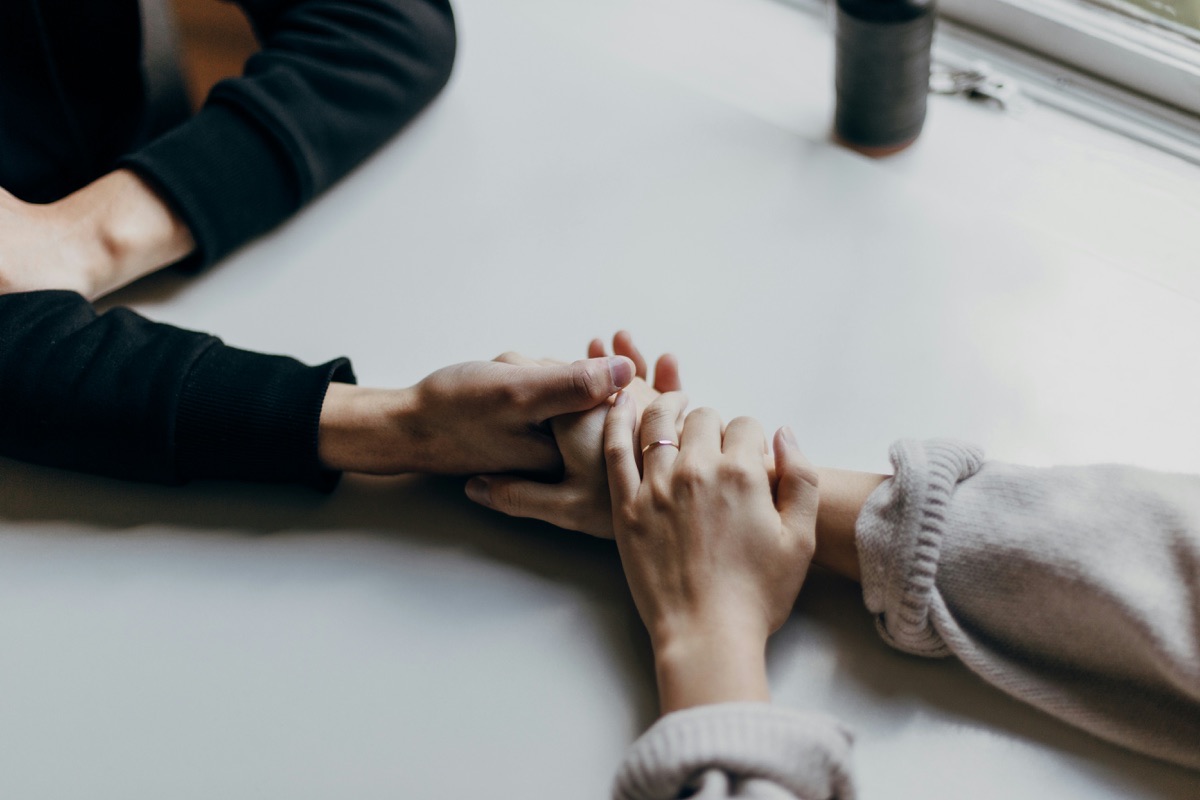 Two people hold hands across a white table; one wears a ring and both wear long sleeves.