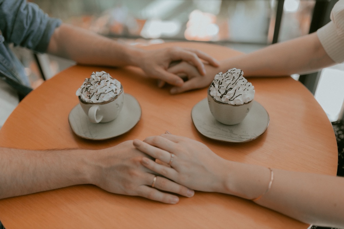 Two pairs of hands rest on a small round table beside two cups topped with whipped cream.