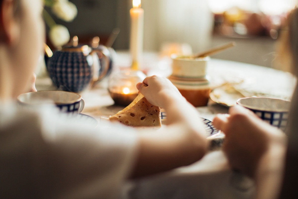 A child's hand holds a folded pancake at a breakfast table set with blue-and-white cups, a teapot and a lit candle.