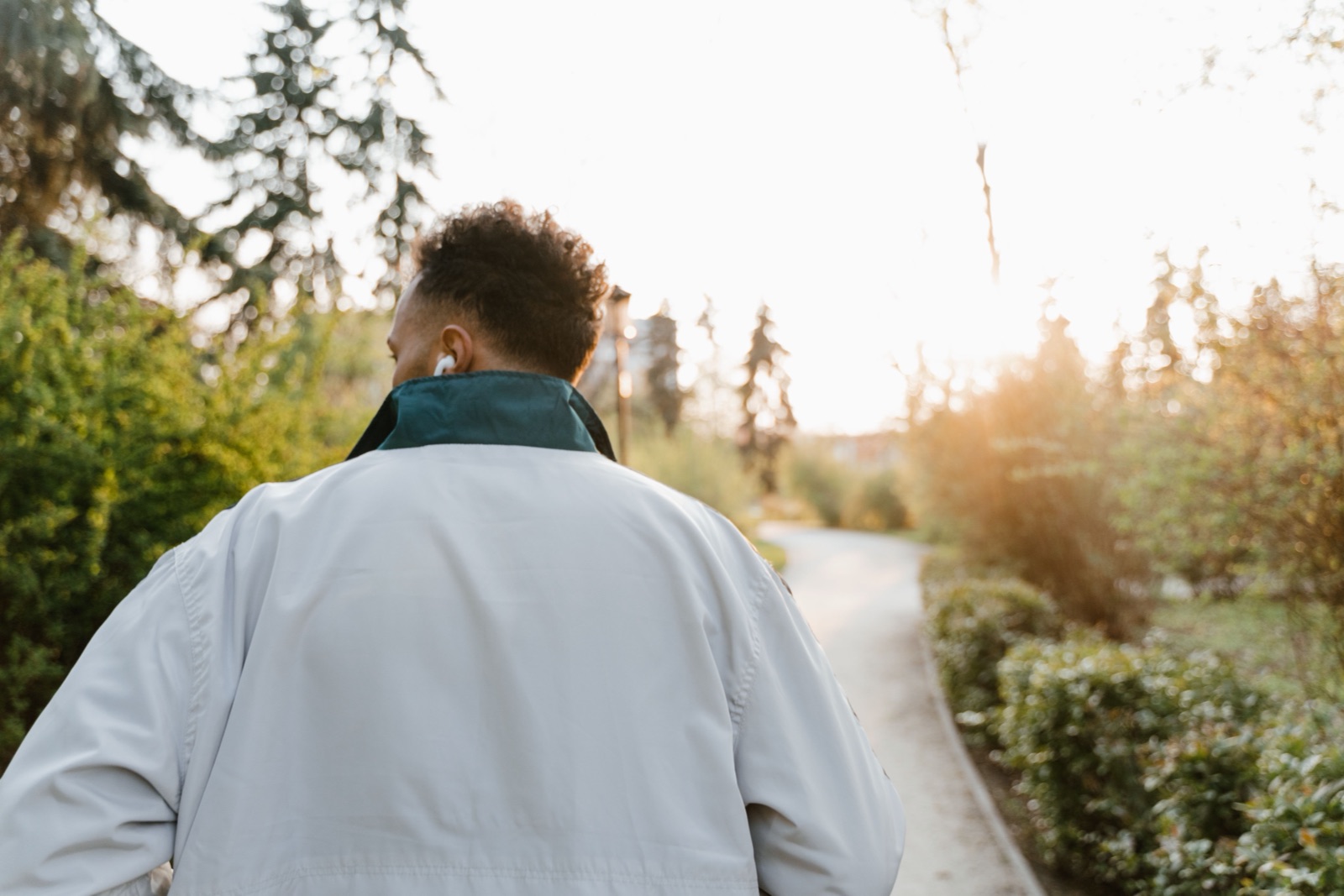 A man in a pale jacket, seen from behind, walks along a park path in low sunlight.