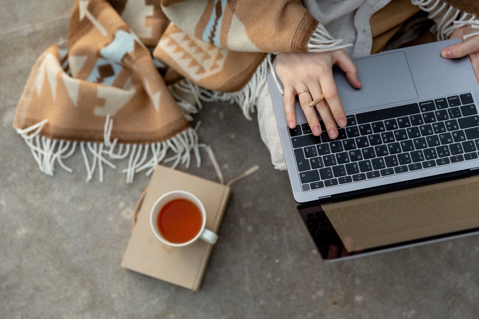Seen from above, a person types on a laptop beside a cup of tea and a patterned blanket.