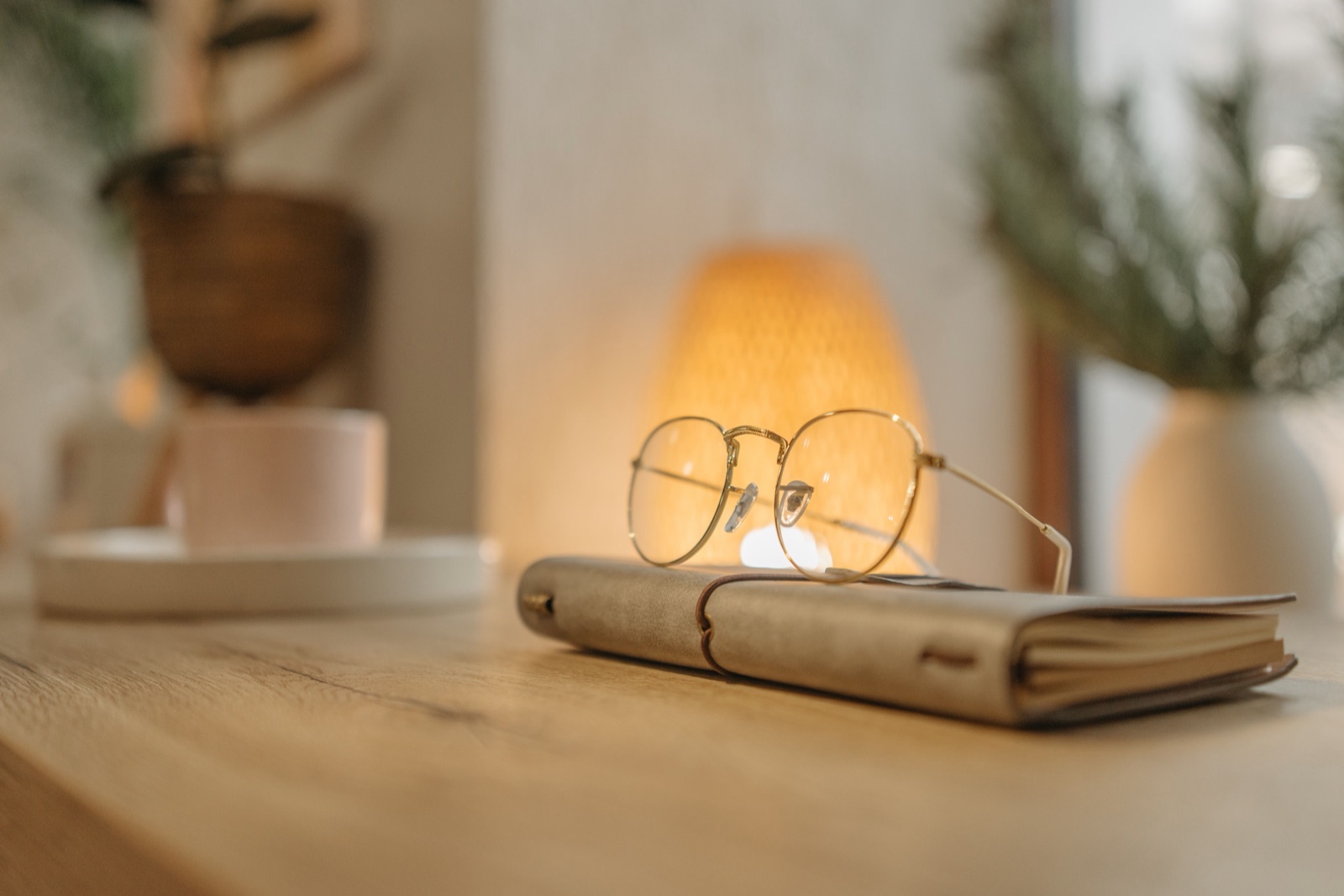 Round wire glasses rest on a leather-bound notebook on a wooden desk, with a warm lamp behind.
