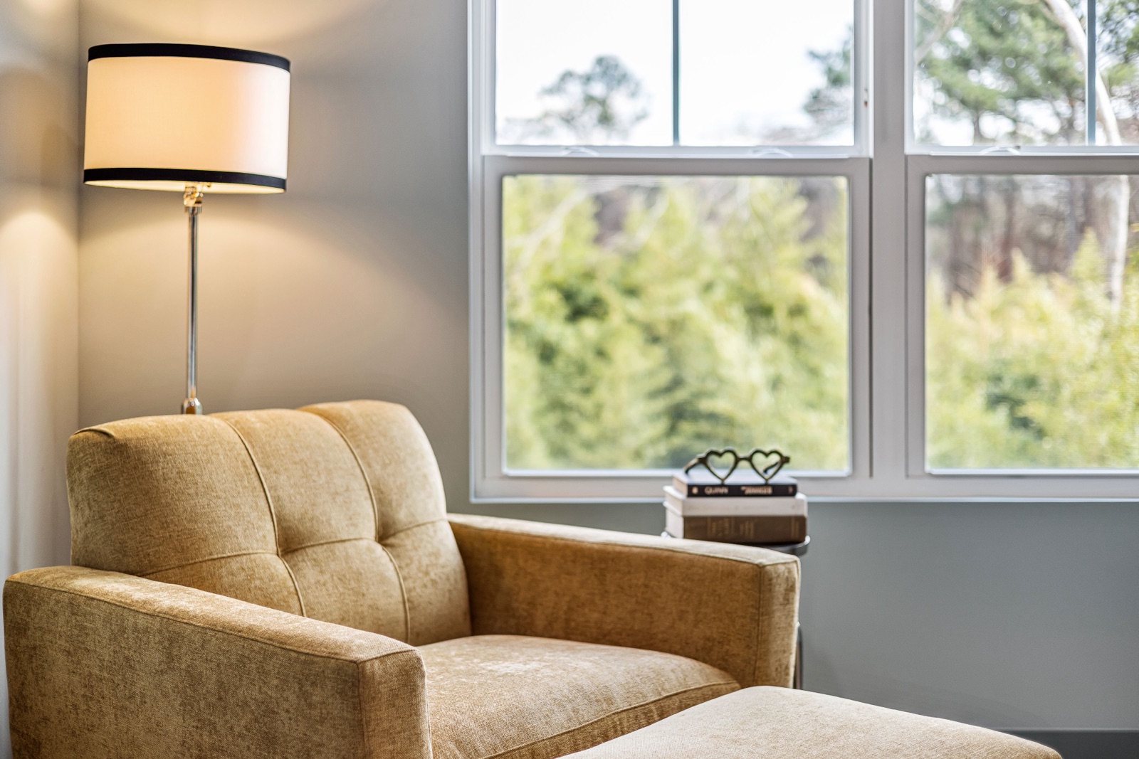 A mustard-coloured armchair and floor lamp sit beside a window looking onto evergreen trees.