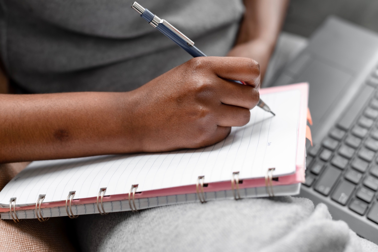 A person writes in a spiral notebook resting on their lap beside a laptop.