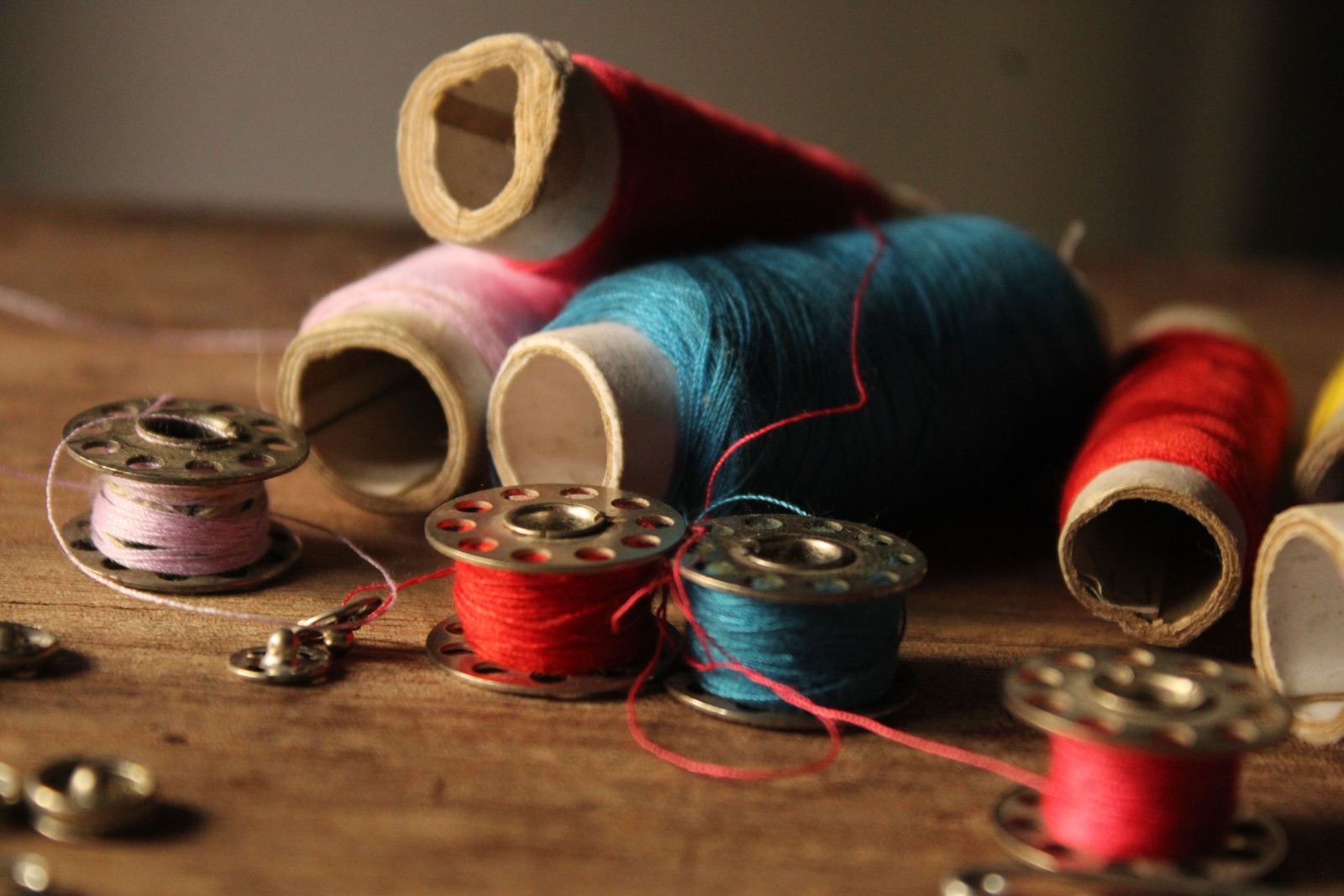 Spools of pink, teal and red thread and metal bobbins lie on a wooden surface.