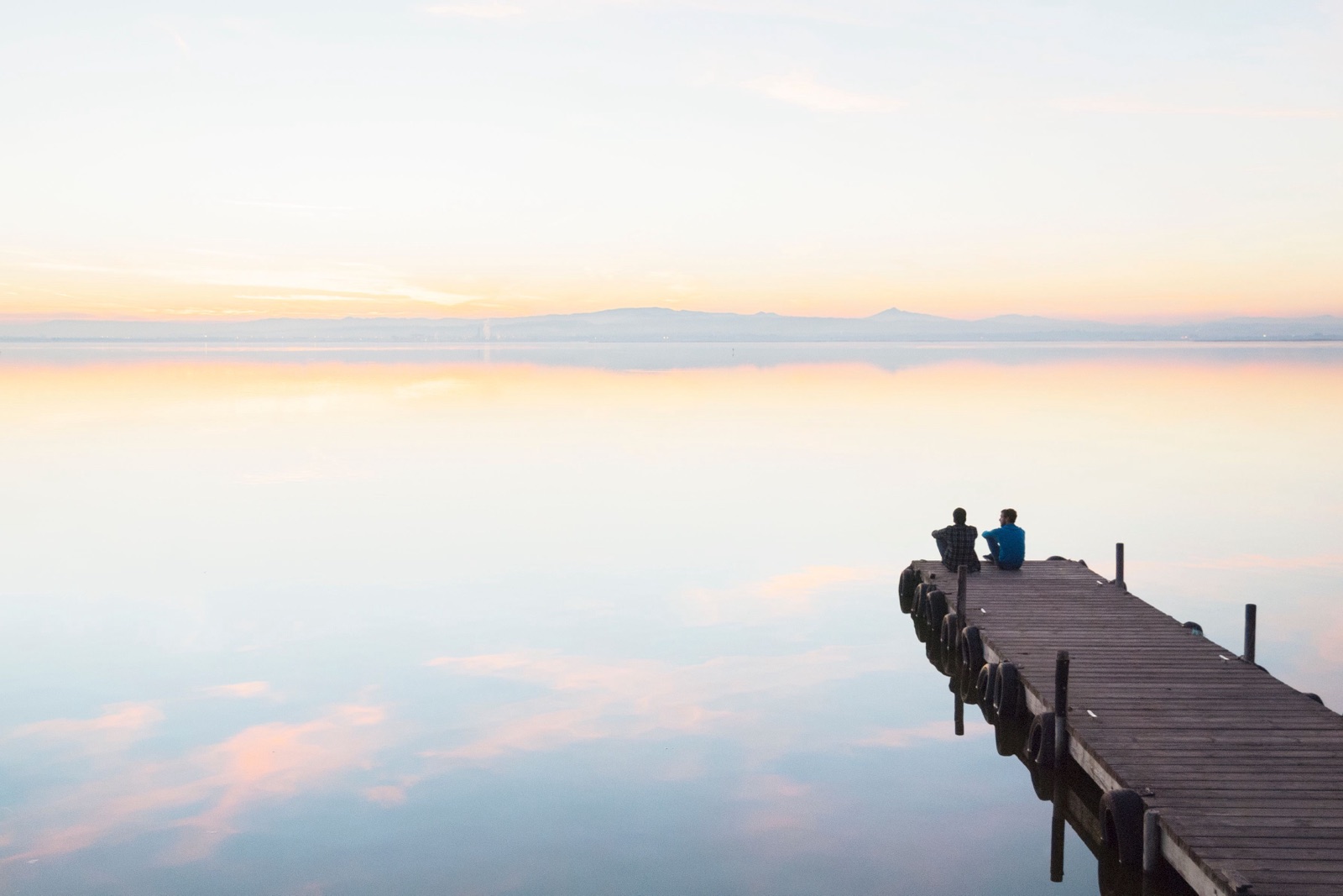 A wooden dock stretches into a still lake under a pale pastel sky, with two people sitting at its end.