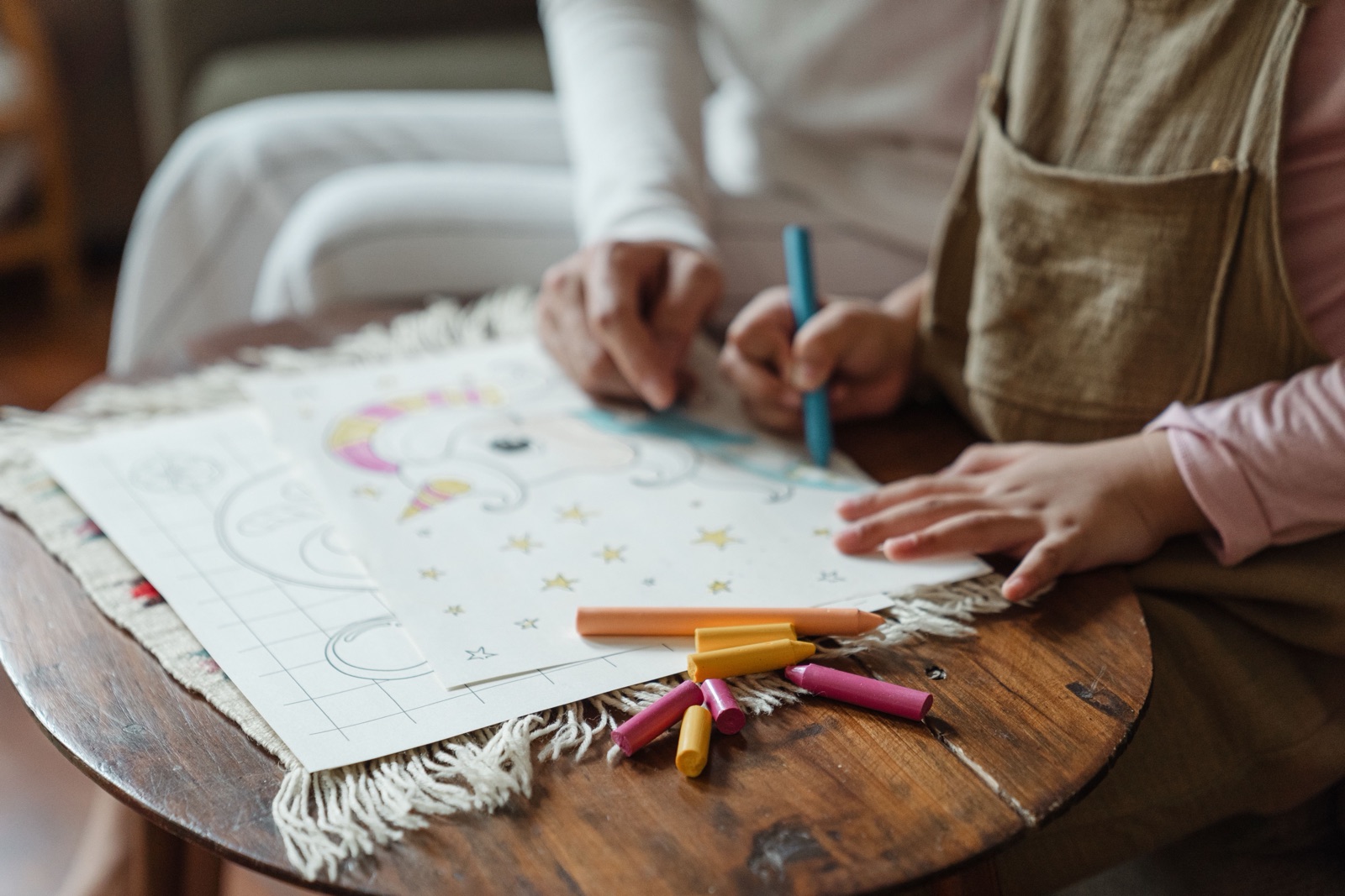 An adult and a small child colour a picture together with crayons at a round wooden table.