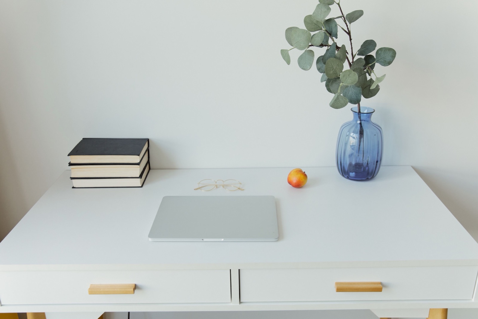 A white desk holds a stack of books, a closed laptop, an apple and a blue vase of eucalyptus.