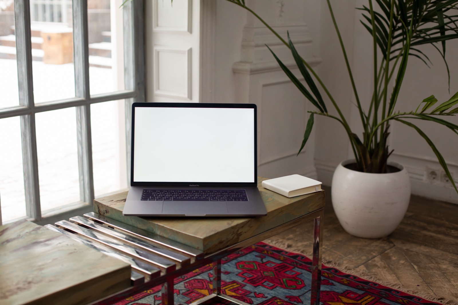 A laptop with a blank screen sits on a low table beside a potted palm and a tall window.