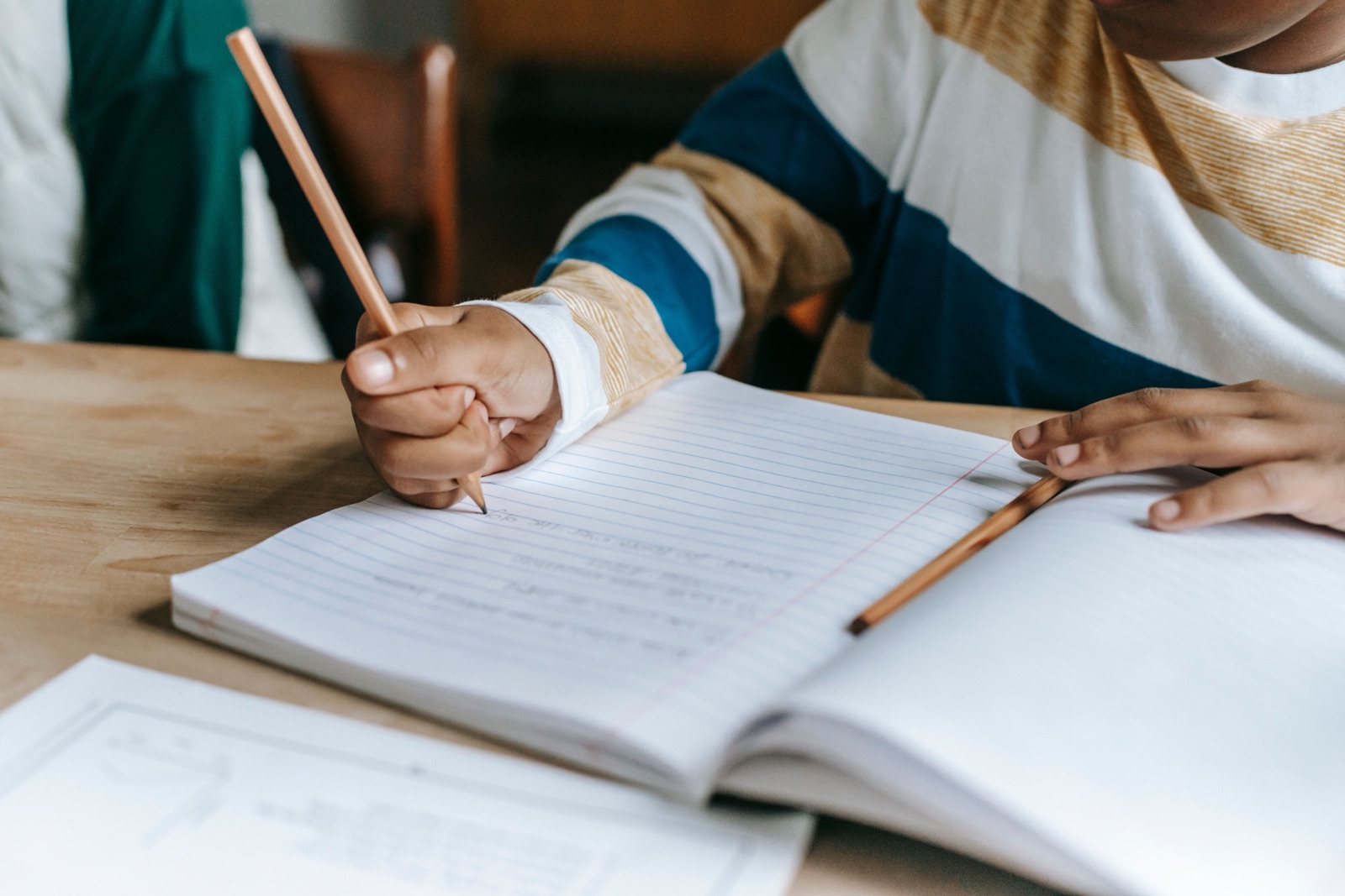 A child in a striped sweater writes with a pencil in a lined notebook at a wooden table.