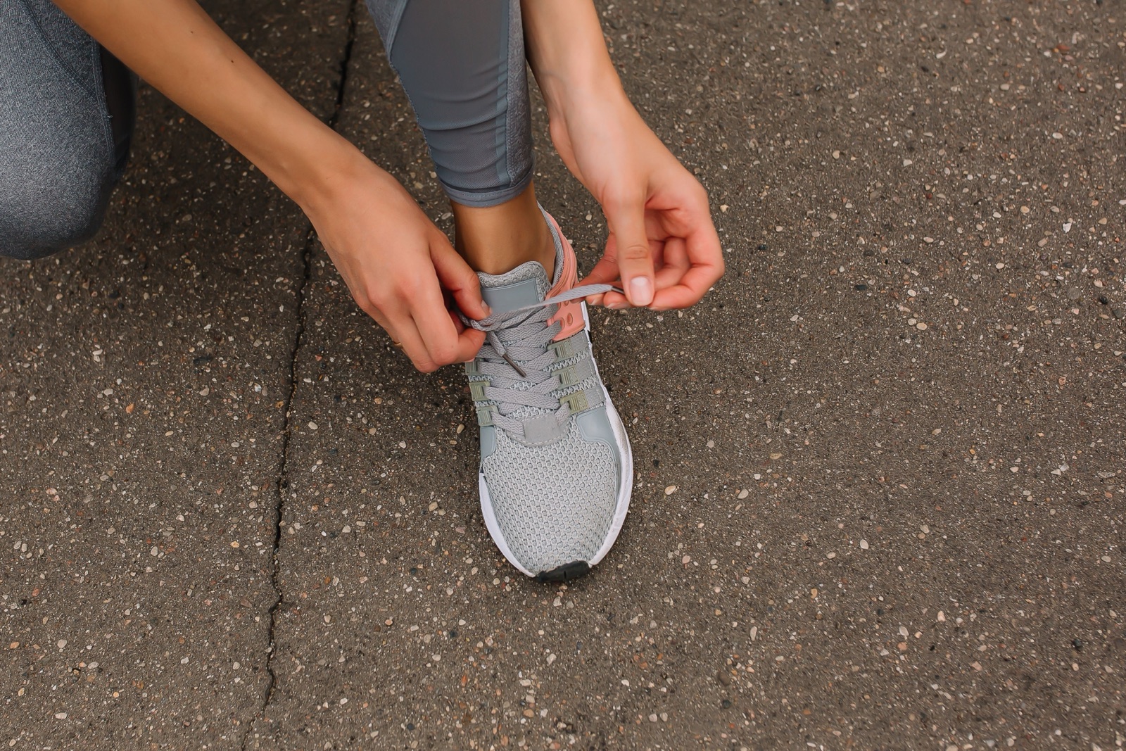 A person crouches on pavement to tie the lace of a grey running shoe.