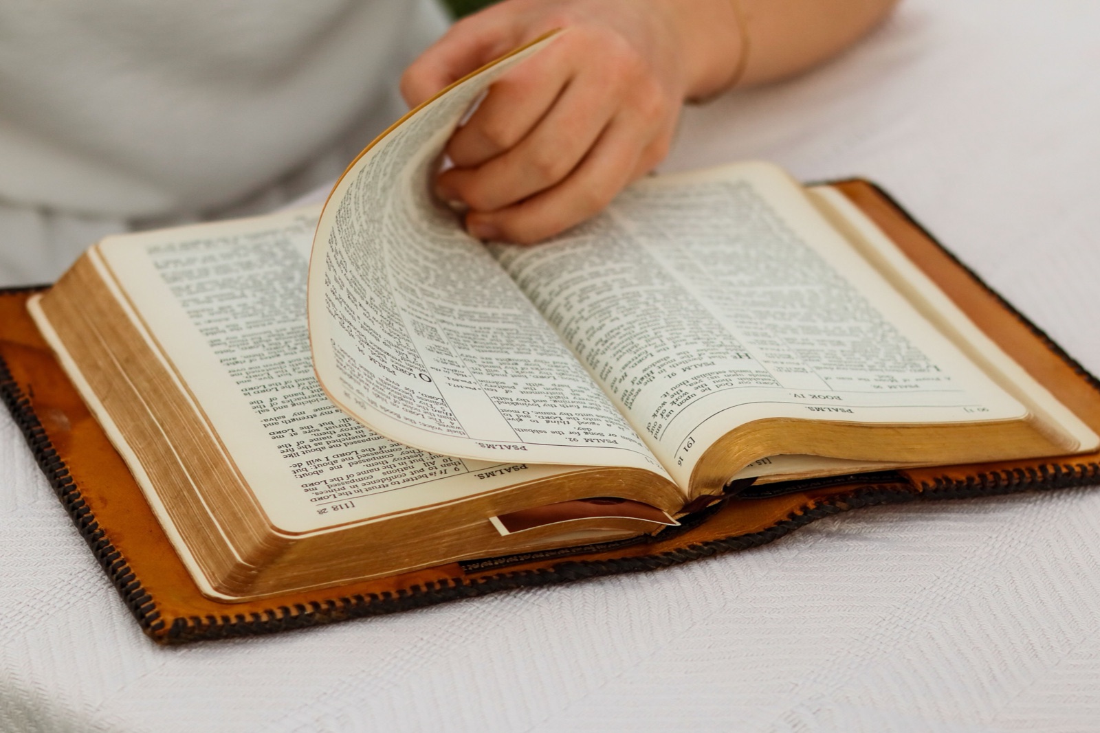 A hand turns a page of an open Bible resting on a white tablecloth in daylight.