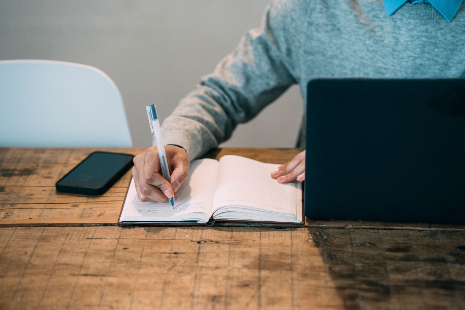 A person in a grey sweater writes in a notebook at a wooden table beside a laptop and phone.