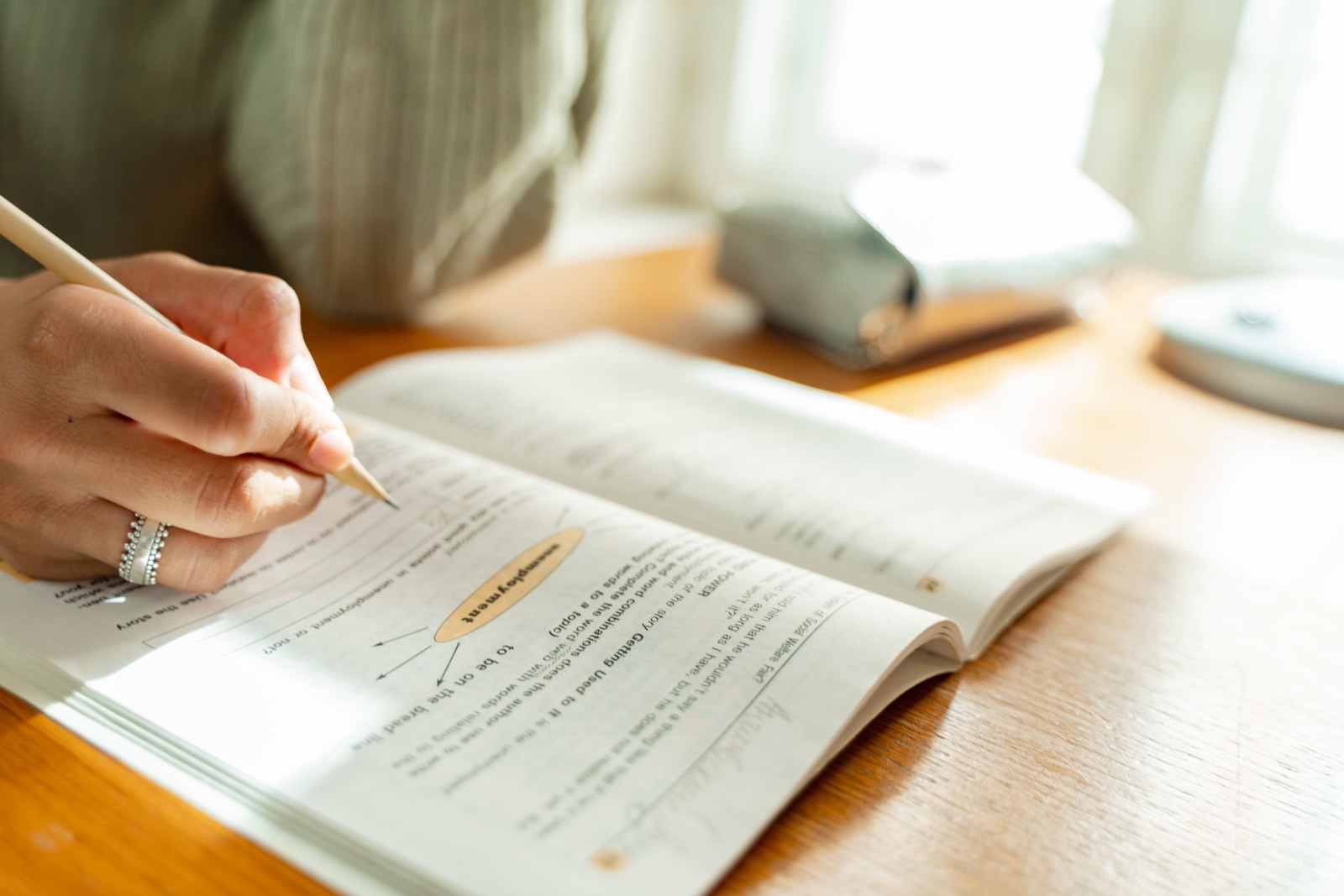 A hand with a pencil works through a printed workbook on a sunlit wooden table.