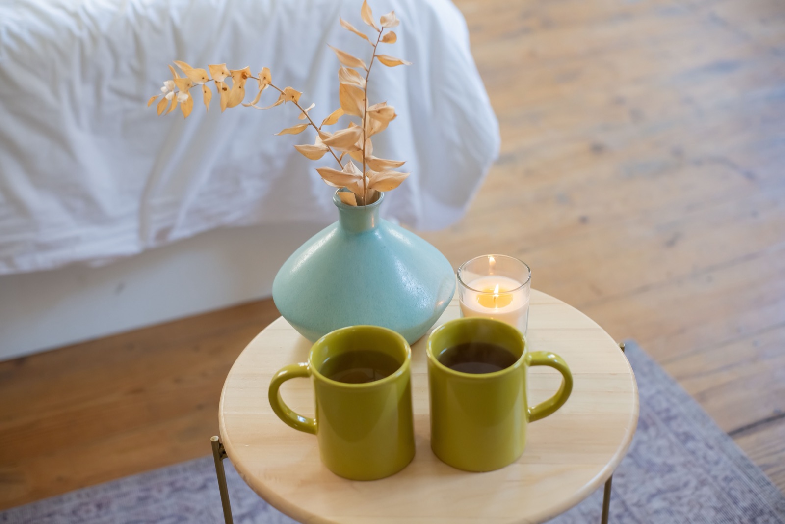 Two green mugs of tea sit side by side on a small round table with a blue vase and a lit candle.