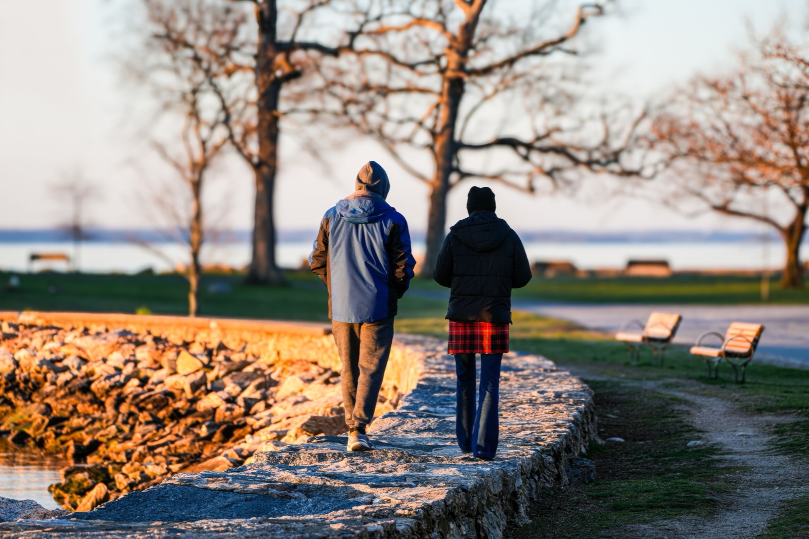 Two people in winter jackets walk side by side along a stone seawall at sunset, seen from behind, with bare trees and water beyond.