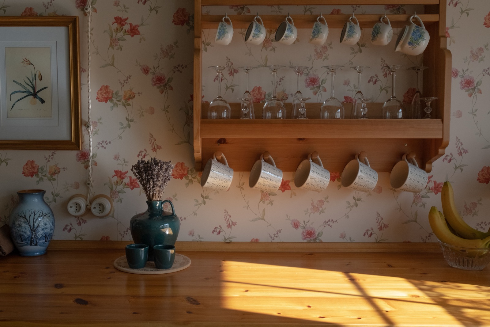 Rows of patterned cups hang from a wooden rack above a pine counter in low sunlight, with a vase of dried lavender below.