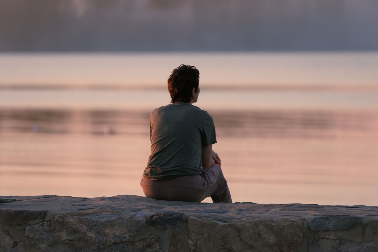 A person seen from behind sits on a stone wall looking over calm water at dusk.