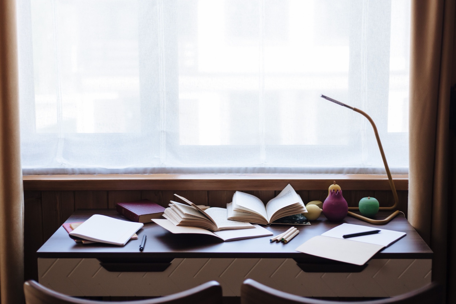 Open books and notebooks are spread on a desk in front of a sheer-curtained window.